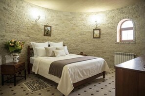 Un dormitorio con pared de piedra, cama con ropa de cama blanca, mesita de noche con un florero y ventana con vistas.