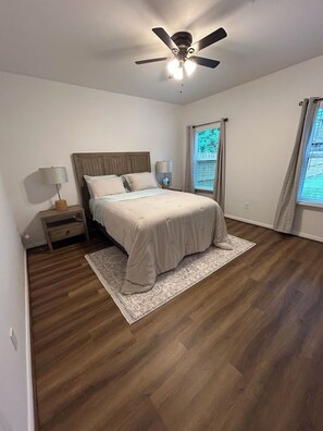 Ein Schlafzimmer mit Bett, Nachttischen, Deckenventilator und einem Fenster mit Vorhängen.