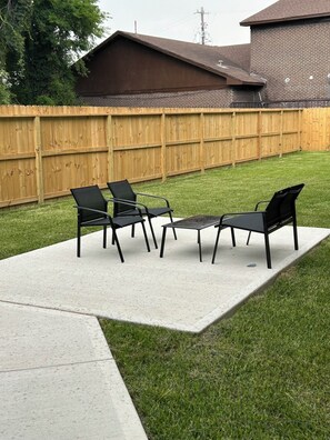 Eine Terrasse mit vier schwarzen Stühlen und einem Tisch, umgeben von einem Holzzaun und einem Rasen.