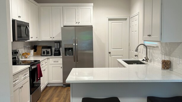 A modern kitchen with white cabinets, stainless steel appliances, a wooden floor, and a large island with two stools. - Phenomenal 3-bedroom townhome with AC in Houston (Houston)