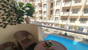 En balkon med fletstole, et rundt bord med en vase og udsigt til en swimmingpool og en høj bygning.
