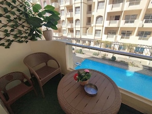 Ein Balkon mit Korbstühlen, einem kleinen runden Tisch mit Blumenvase und Blick auf ein Schwimmbecken sowie ein mehrgeschossiges Gebäude.