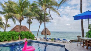 A pool with a pink flamingo float, palm trees, a thatched-roof hut, and a view of the ocean. -  Charming Casita Beach and Pool Access 2 king beds (Q.R.)