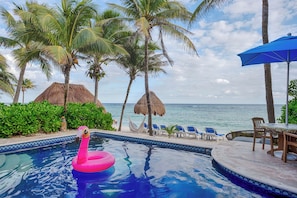 Ein Pool mit einem Flamingo-Schwimmding, Palmen, einer Hütte mit Reetdach und Blick auf den Ozean.