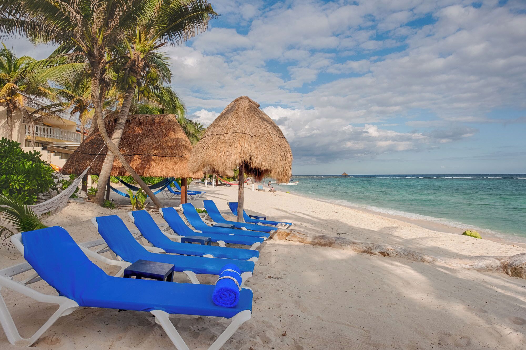 Una playa con sombrillas de techo de paja, sillas reclinables azules y palmeras.