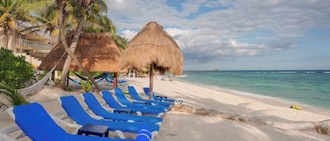 A beach with thatched umbrellas, blue lounge chairs, and palm trees.
