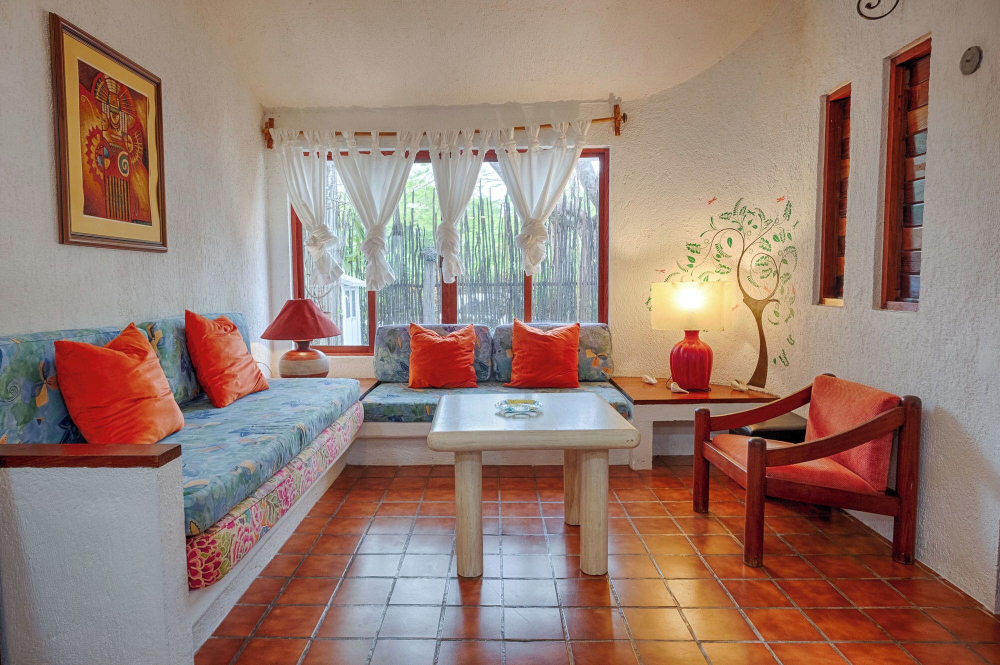 Una sala de estar acogedora con un sofá, una mesita y un sillón. Se ve una ventana con cortinas, una lámpara y un adorno en la pared.