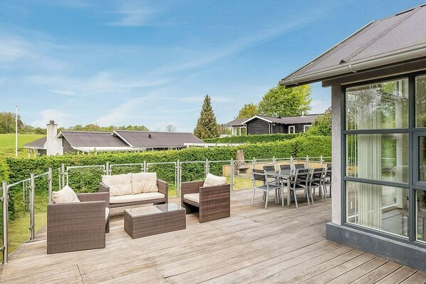 Una terraza con mobiliario de mimbre, una sala con paredes de cristal y vistas a casas al fondo.