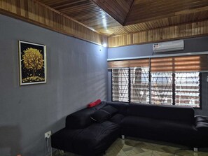 Ein Zimmer mit Holzboden, einem schwarzen Ecksofa, einem gerahmten Bild an der Wand und einem Fenster mit Jalousien.