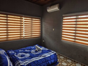 Ein Schlafzimmer mit einem Bett, zwei Fenstern mit Jalousien, einer Klimaanlage und einem gemusterten Boden.