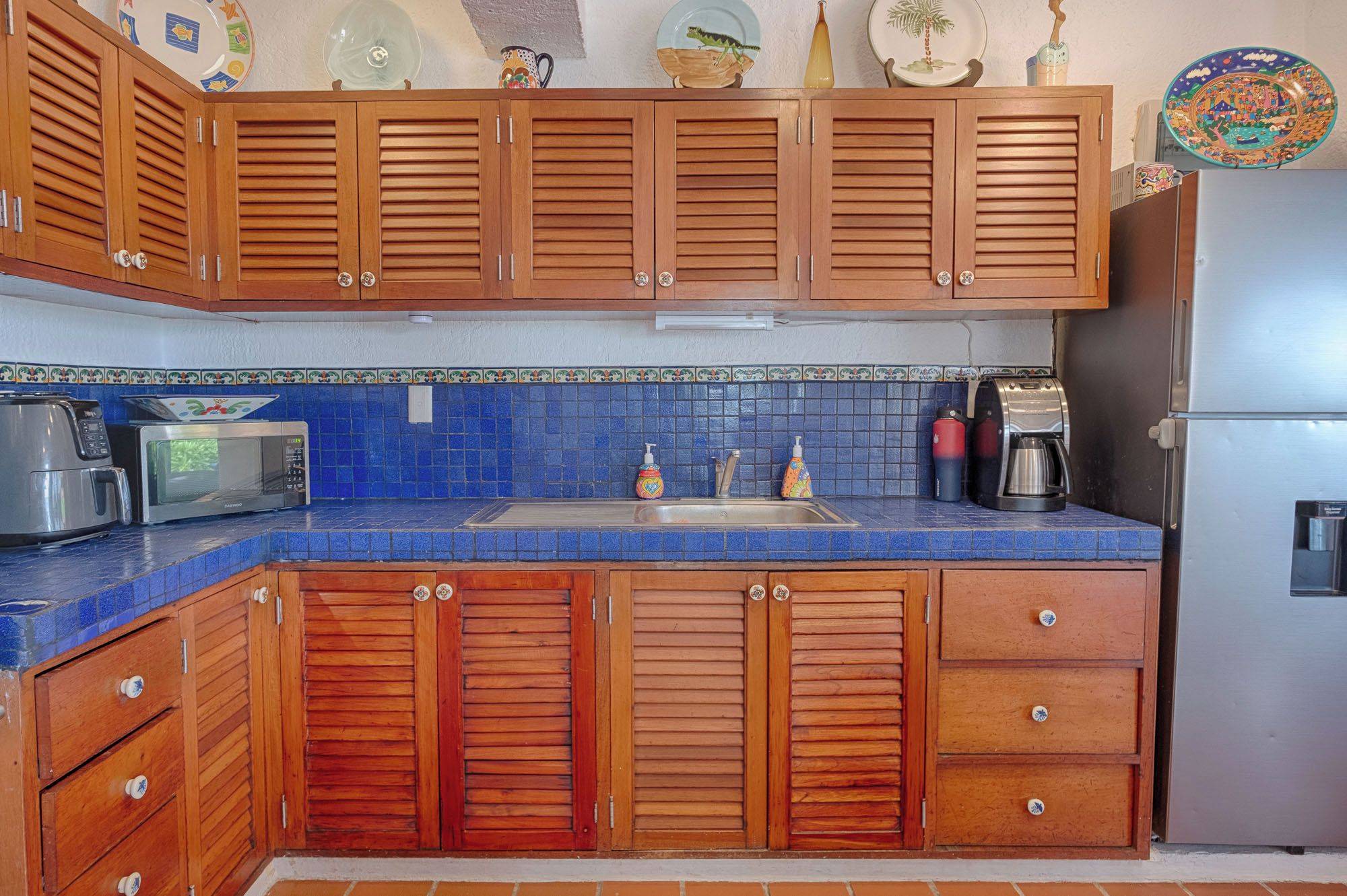 Een keuken met houten kastjes, een blauwe betegelde spatwand, en diverse apparaten, waaronder een magnetron en koffiezetapparaat.