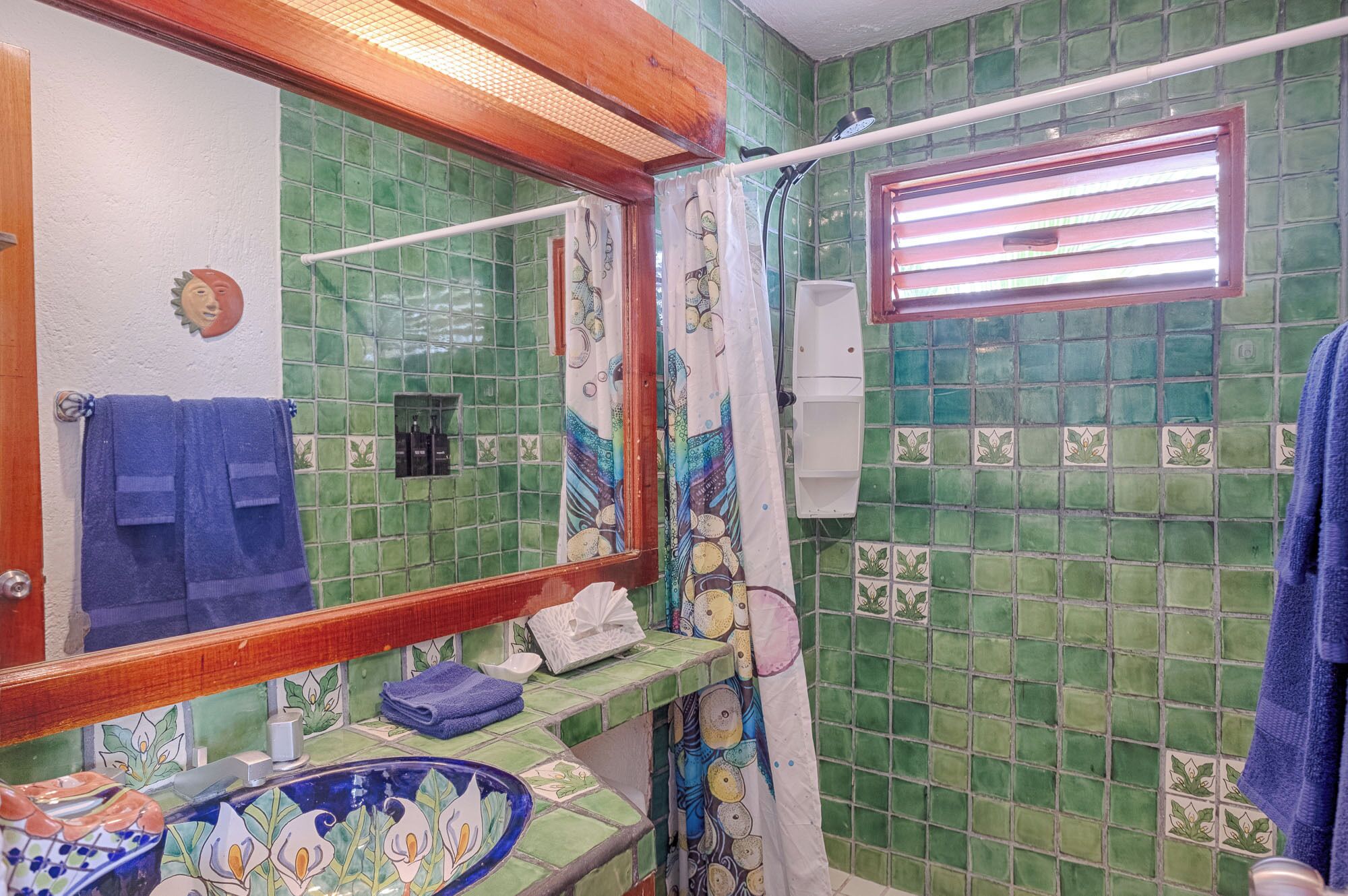 Un baño con paredes de azulejos verdes, un lavamanos azul con motivos florales, un espejo con marco de madera y una ducha con cortina.