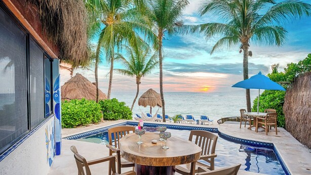 Un espace aménagé au bord de la piscine, avec une table ronde en bois et des chaises, entouré de palmiers et offrant une vue sur l’océan au coucher du soleil.