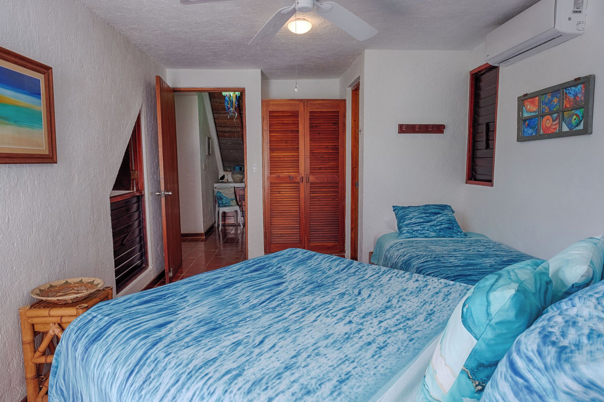 Une chambre avec deux lits, une porte en bois, un ventilateur de plafond et un tableau accroché au mur.