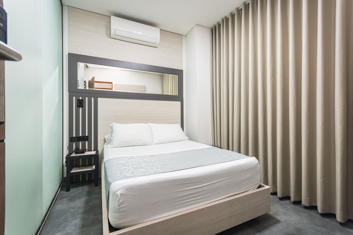 A hotel room with a bed, a bedside table, a mirror, a wall-mounted air conditioner, and a window with curtains.