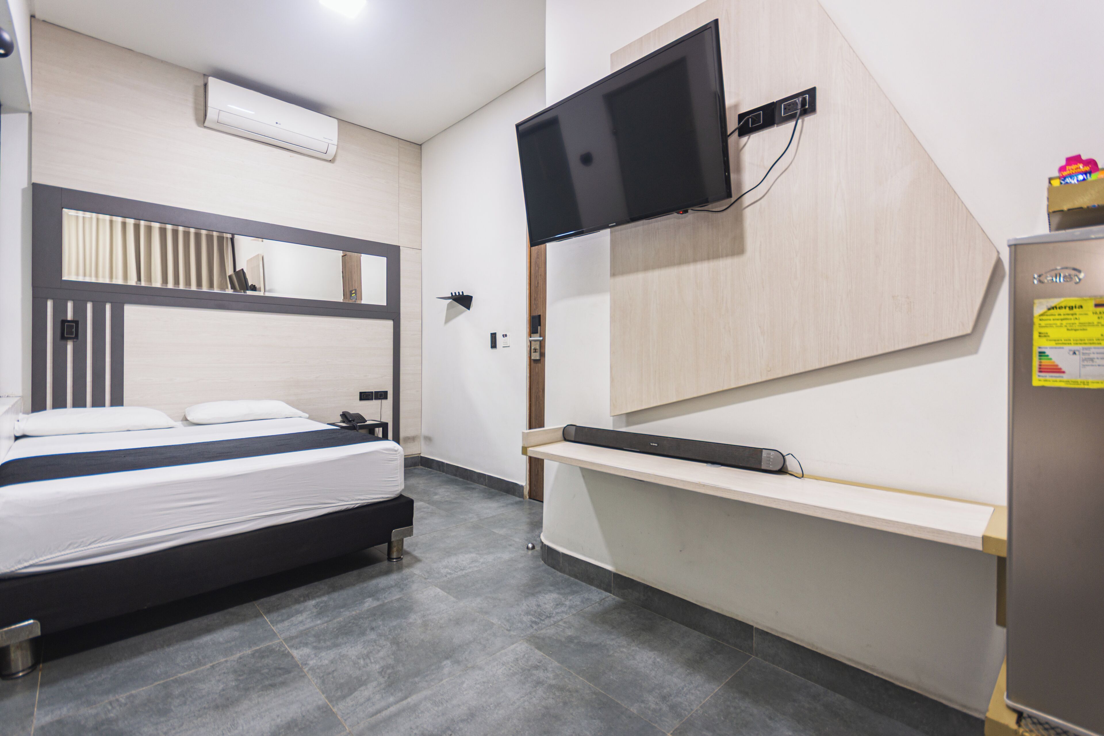 A modern hotel room with a bed, a mounted TV, a built-in shelf, and a refrigerator.