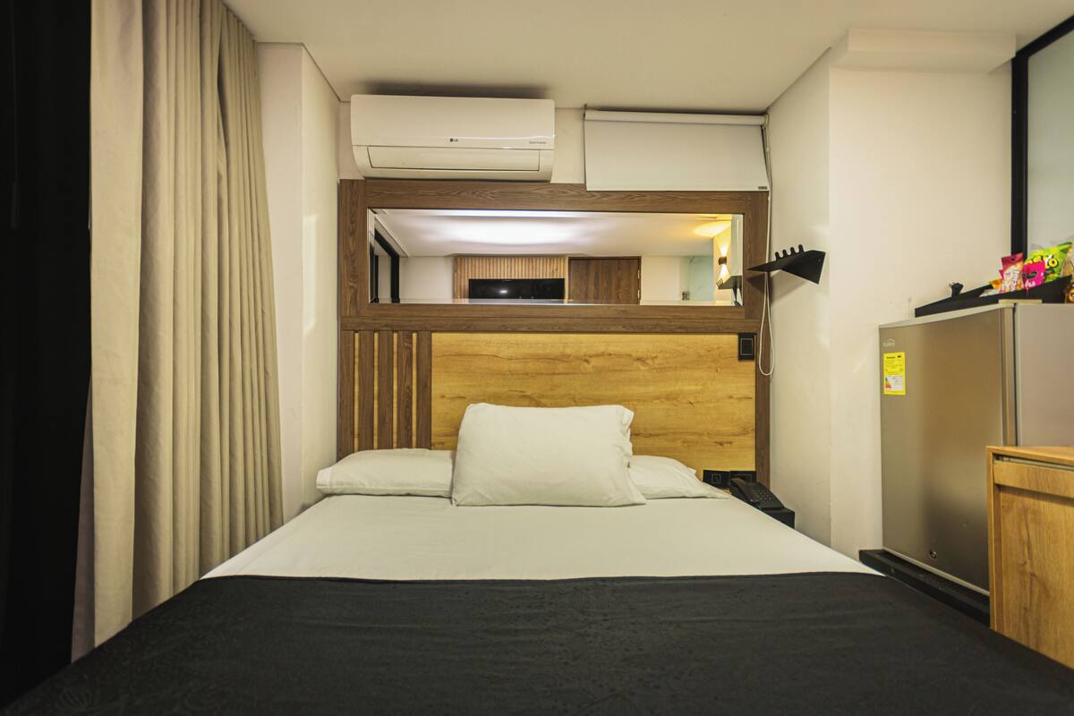 Una habitación de hotel con una cama, cabecera de madera, espejo, equipo de aire acondicionado y refrigerador.