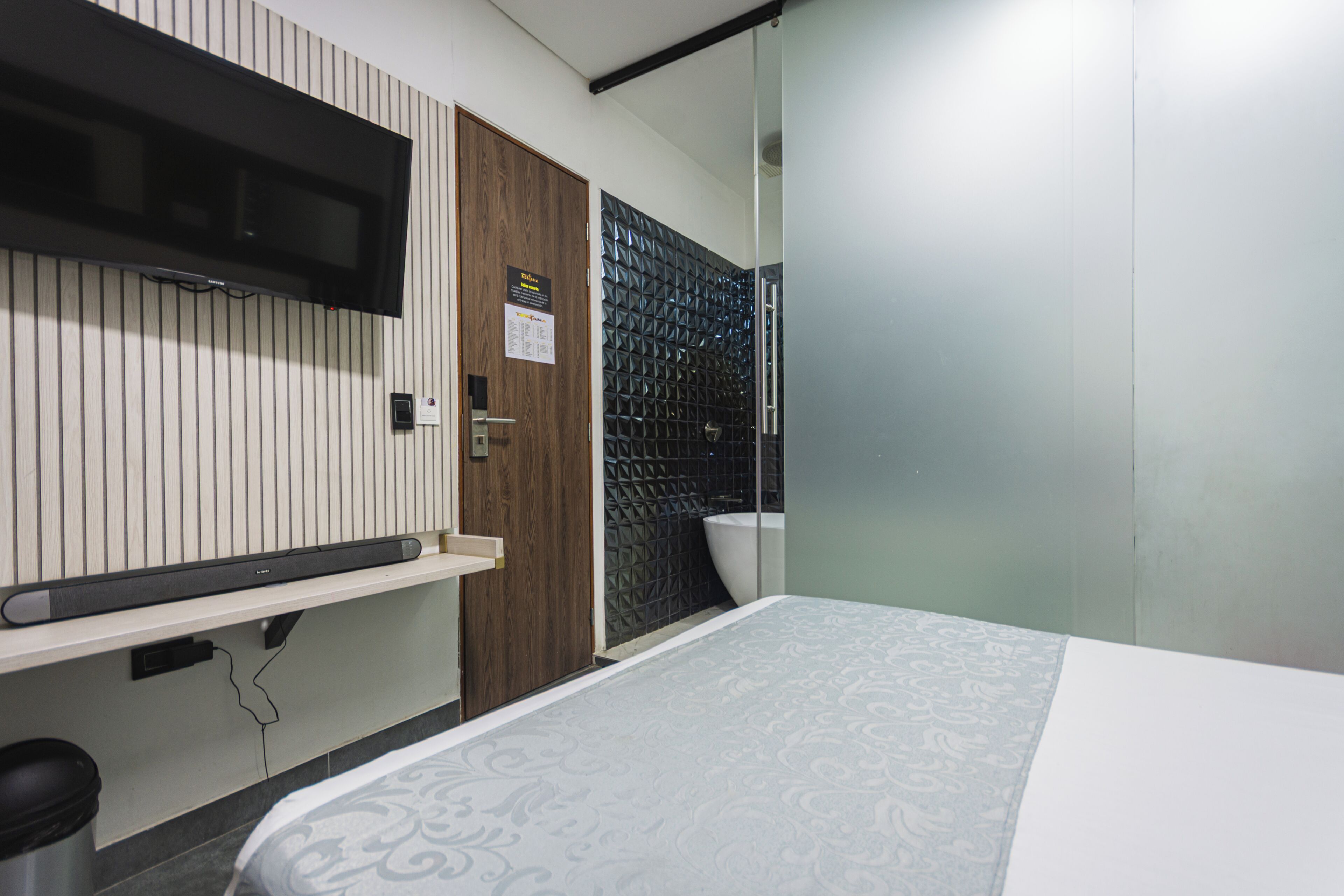 Habitación de hotel con cama, televisión montada en la pared, puerta de madera y zona de ducha con azulejos.