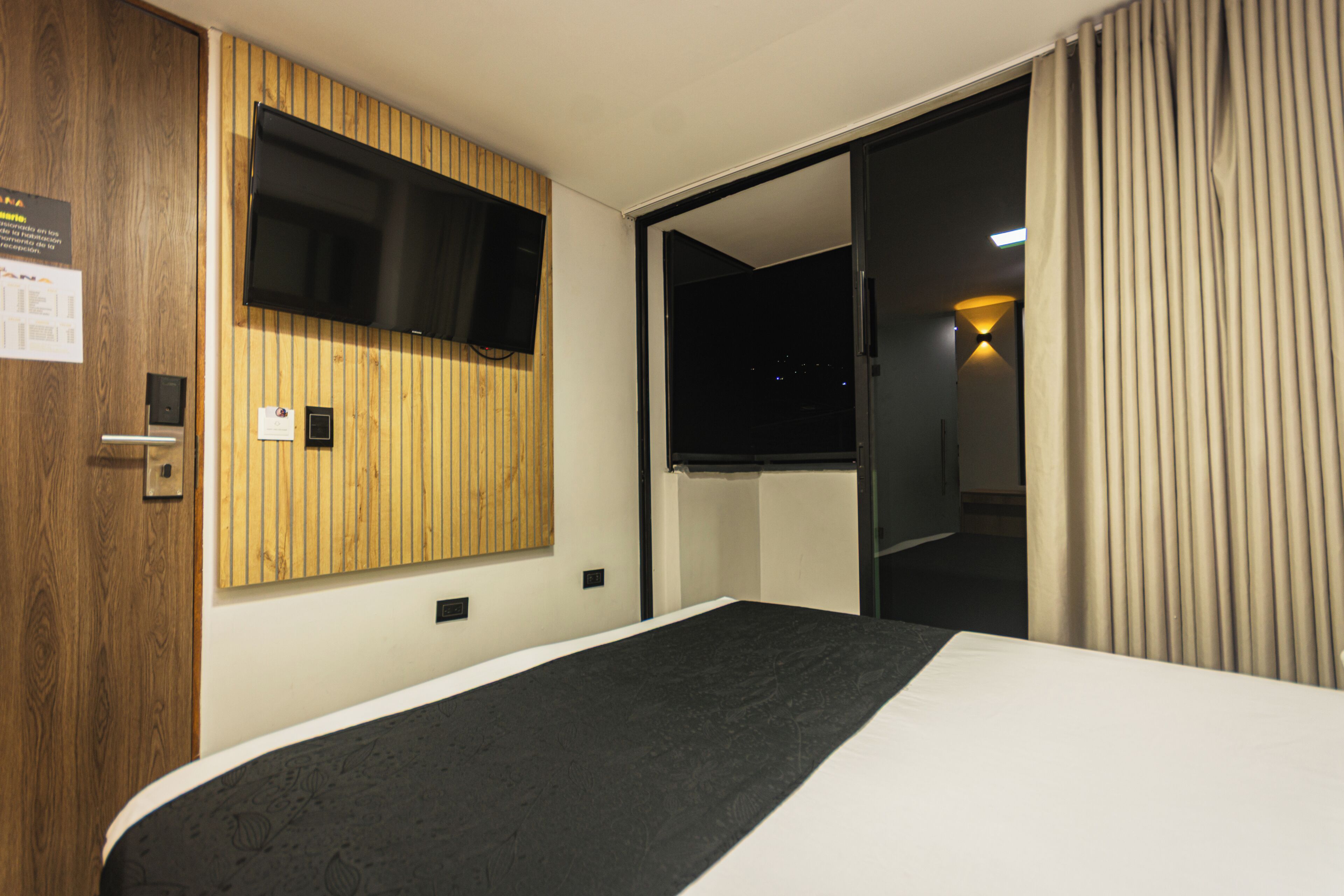 A hotel room with a bed, a TV mounted on a wooden panel wall, a door, and a curtain.