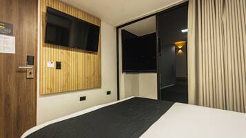 A hotel room with a bed, a TV mounted on a wooden panel wall, a door, and a curtain.