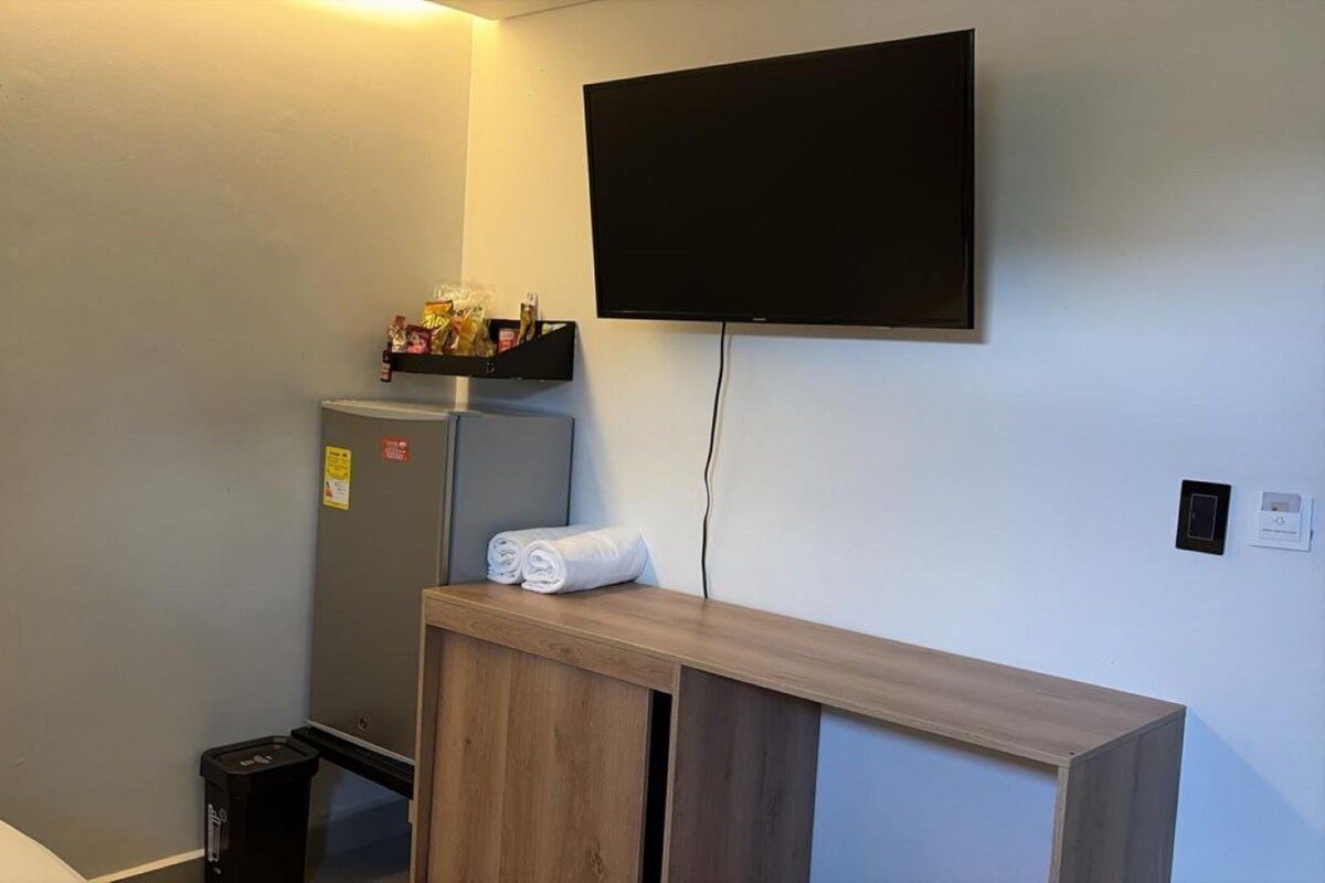 Habitación compacta con televisión integrada, un refrigerador pequeño y una consola de madera con toallas.