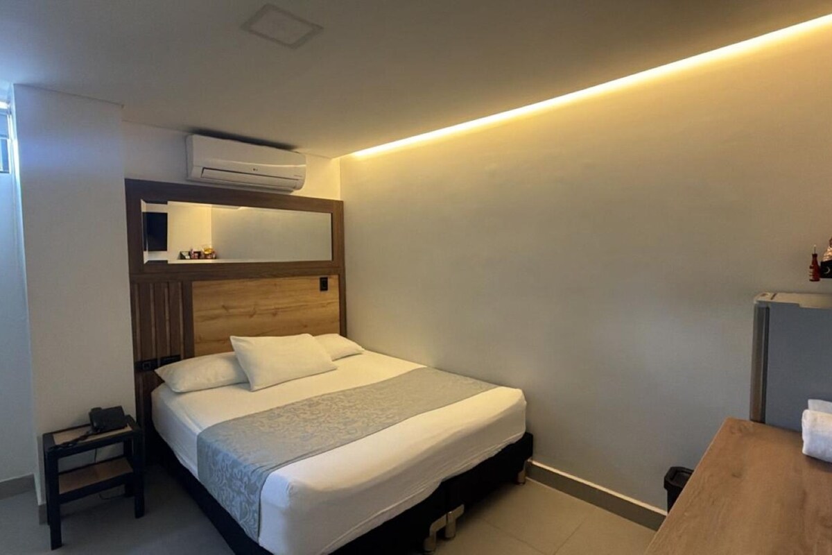 Une chambre d’hôtel comprenant un lit, une table de chevet, une petite commode, un climatiseur fixé au mur et une étagère avec des articles de toilette.