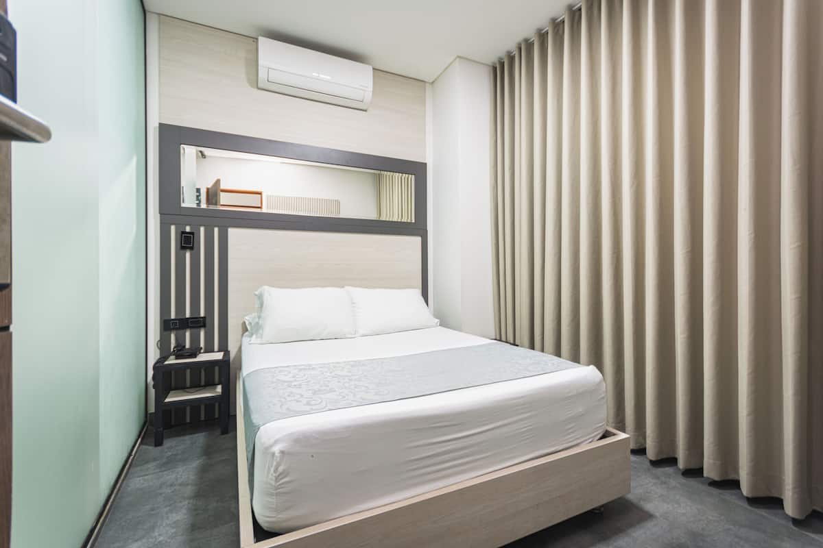 Una habitación de hotel con cama, mesita de noche, aire acondicionado y cortinas.