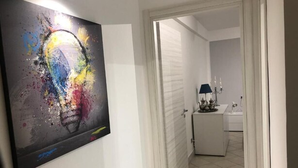 Un pasillo con un cuadro abstracto colorido en la pared, una puerta blanca y una vista parcial de un baño con un lavabo y una lámpara.
