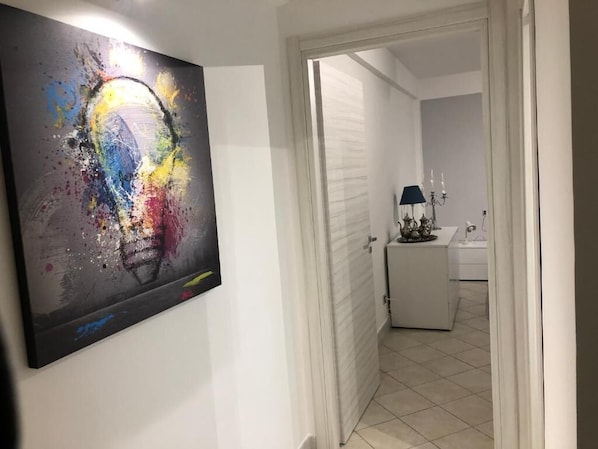 Ein Flur mit einem farbenfrohen, abstrakten Gemälde an der Wand, einer weißen Tür und einem Blick ins Badezimmer mit Waschbecken und Lampe.
