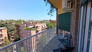 Un balcon avec une table et des chaises, un volet roulant vert et une vue sur les bâtiments environnants.