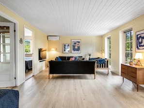 Ein geräumiges Wohnzimmer mit einer großen schwarzen Couch, einem Holztisch und einem an der Wand befestigten Fernseher. Zwei Fenster sorgen für natürliches Licht, und eine Tür führt in einen anderen Raum.