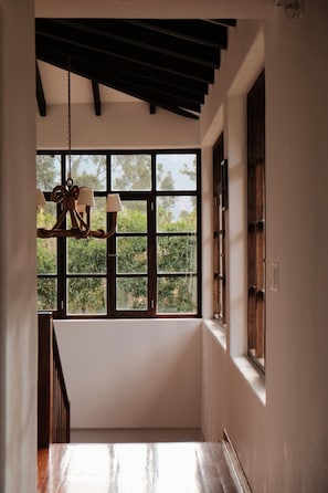 A hallway with a wooden staircase, a hanging chandelier, and a large window with a view of greenery.