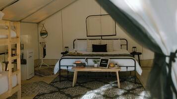 A tented accommodation room with a bed, bedside table, and a small table with a sign.