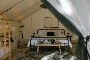 A tented accommodation room with a bed, bedside table, and a small table with a sign.