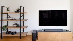 A modern living room with a TV mounted on the wall, a wooden entertainment unit, and a metal shelving unit with model cars and plants.