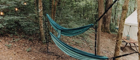 Una hamaca colgando entre árboles en un área boscosa, con una tienda de campaña y una mesa de picnic cerca.