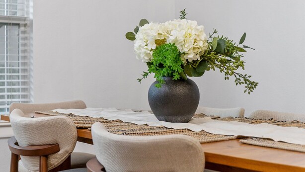 Une table à manger agrémentée d’un vase de fleurs blanches, de sets de table en fibres tressées et de chaises modernes.