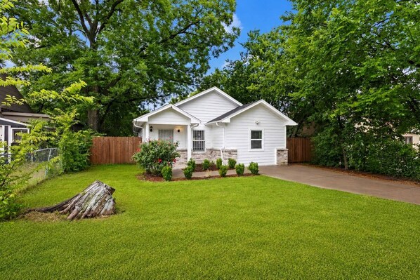 A single-story house with a front porch, a small front yard with a wooden fence, and a driveway leading to the garage. - Modern 4BHK Backyard 25 mins FIFA 10 mins Downtown (Dallas)