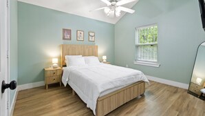 Une chambre à coucher comprenant un lit, des lampes de chevet, un ventilateur de plafond, une fenêtre avec des stores et des œuvres d’art encadrées accrochées au mur.