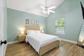 Ein Schlafzimmer mit Bett, Nachttischlampen, Deckenventilator, einem Fenster mit Jalousien und gerahmten Kunstwerken an der Wand.