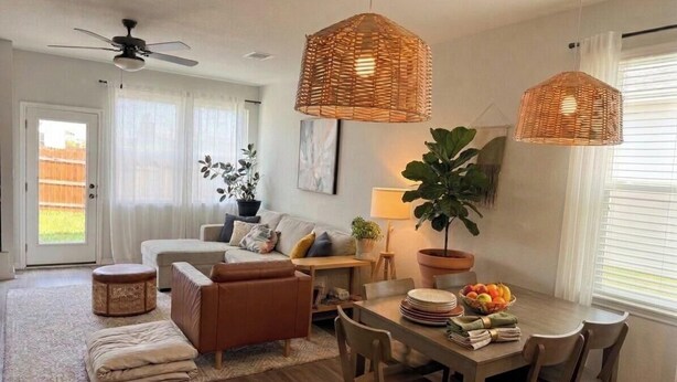 En moderne stue med sofa, sofabord og spisebord. Der er fletlamper, en loftsvifte og en stor plante.