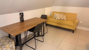 Ein kleiner Raum mit einem Holztisch, zwei Stühlen, einem gelben Sofa und einem gemusterten Kissen.