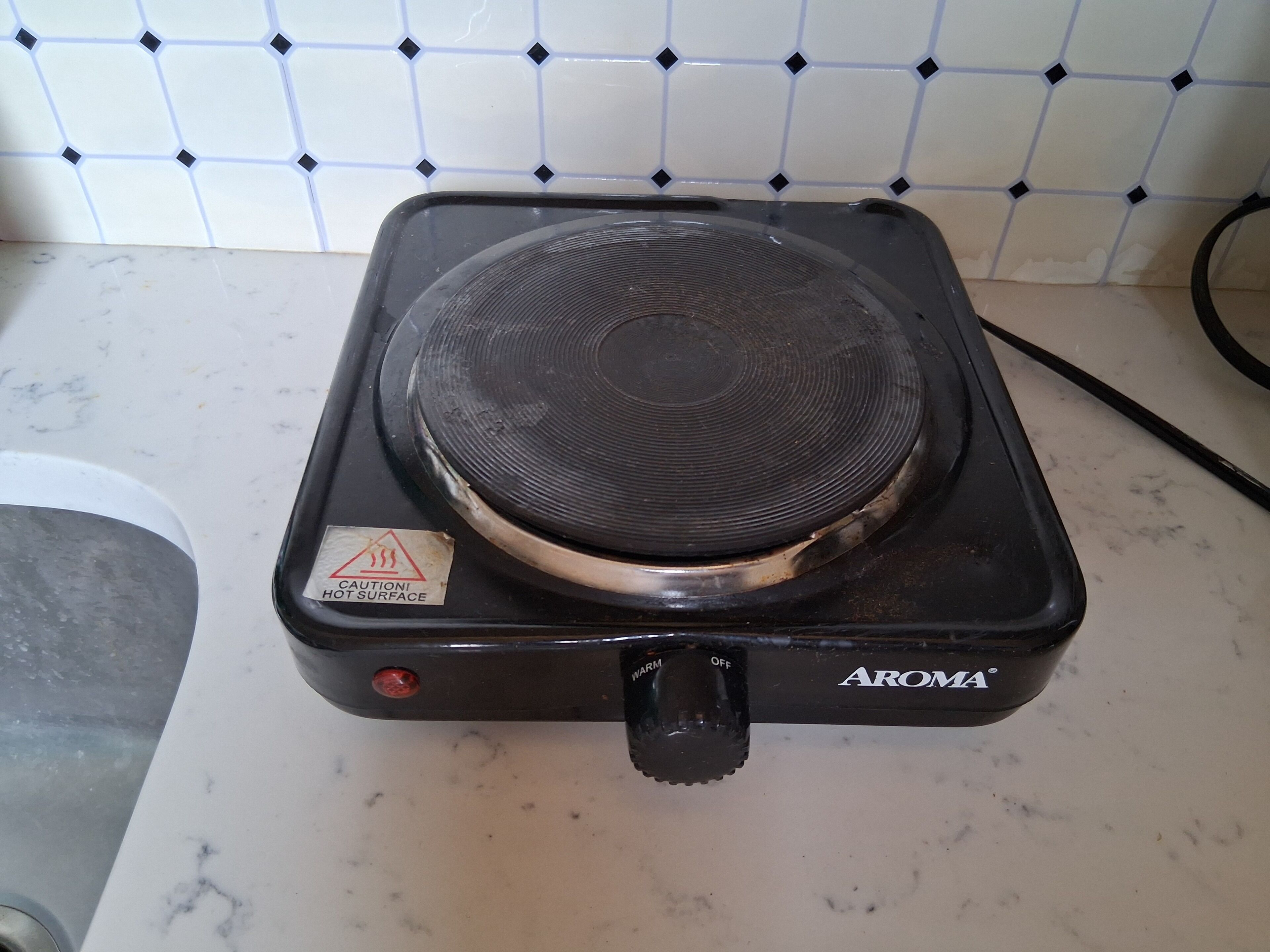 Um fogão elétrico preto com um aviso de segurança, um botão de controle e a marca 'AROMA' em uma bancada de cozinha.