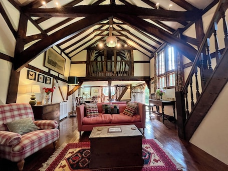 A room with exposed wooden beams, a red sofa, a plaid armchair, and a wooden coffee table.. A unique Suffolk timber-framed cottage set within private lawns and woodland.