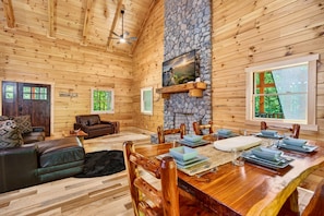 L’intérieur d’un chalet en bois comprend une table à manger pour quatre personnes, une cheminée et une grande fenêtre donnant sur un paysage verdoyant.