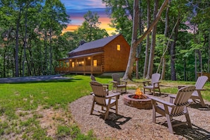 Une cabane en bois avec un porche, entourée d’arbres, et un espace extérieur aménagé avec un foyer.