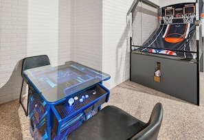 A room with a glass-top table, a black chair, a basketball arcade machine, and a white brick wall. - Happy Hours Cabin/Hot Tub & Hocking Hills (Logan)