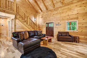 Ein Interieur einer Holzhütte mit einer Holztreppe, Ledersofas, einem Holztisch und einem Fenster mit Blick auf Bäume.