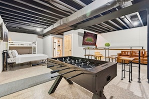 A room with a foosball table, a bed, a sofa, and a television displaying a football match. - Happy Hours Cabin/Hot Tub & Hocking Hills (Logan)