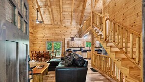 A log cabin interior with a dining area, a sofa, a staircase, and a kitchen. - Tranquil Happy Hours Cabin: Hot Tub, games, fire pit & wooded privacy (Logan)